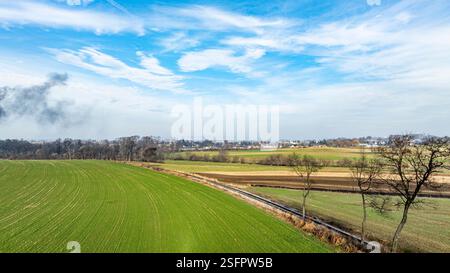 De vastes champs verts s'étendent sous un ciel bleu vif, orné de quelques nuages. À proximité, les forêts et les terres agricoles lointaines mettent en valeur les teintes automnales, créant un paysage rural serein. Banque D'Images