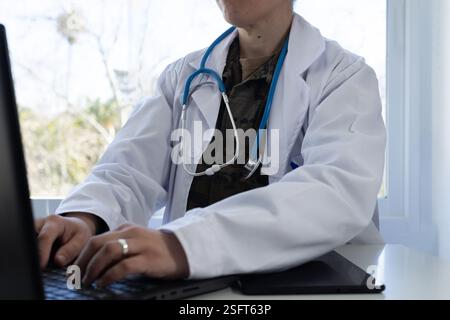 Médecin militaire en manteau blanc et stéthoscope, tapant sur un ordinateur portable dans un bureau bien éclairé. Banque D'Images