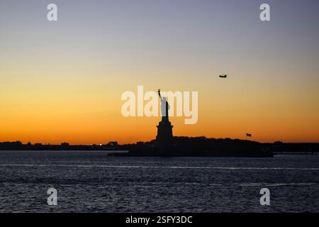 La Statue de la liberté brille au coucher du soleil tandis qu'un hélicoptère Chinook de l'armée américaine vole au-dessus de lui, créant une scène dramatique et patriotique. Banque D'Images