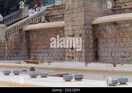 Un mur de pierre avec un aspect altéré, avec un bassin de fontaine au premier plan. Le mur est fait de grosses pierres irrégulières, et il y a des rem Banque D'Images