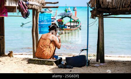 Phangnga, Thaïlande, 21 janvier 2025, Une femme moken se brosse les dents sur la plage de l’île de Surin, des bateaux de pêche colorés flottent doucement à proximité, renforçant l’atmosphère sereine de ce village authentique Banque D'Images