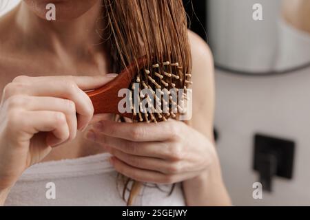 Gros plan d'une femme se brossant les cheveux mouillés avec une brosse à palette en bois. Routine de soins capillaires, beauté et concept d'auto-soin dans un cadre de salle de bain moderne Banque D'Images