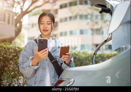 Une belle femme asiatique positive vérifie son passeport, les détails de son vol et son heure d'embarquement sur son smartphone avant de monter dans un taxi pour l'aéroport. Banque D'Images