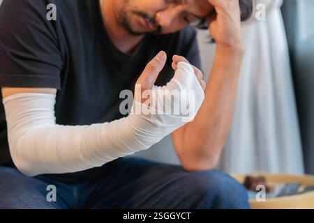 Homme asiatique adulte avec un bandage de gaze enroulé autour de ses bras blessés assis à la maison avec le visage de préoccupation. Mise au point sélective. Banque D'Images