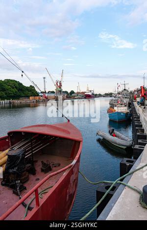Une vue large d'un port industriel avec divers bateaux, dont un rouge proéminent au premier plan, amarré aux côtés de grues et d'autres ports infras Banque D'Images
