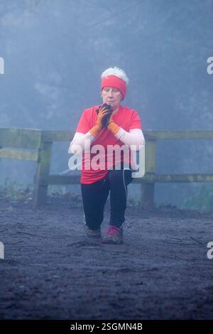 Femme âgée en vêtements de sport faisant des fentes à l'extérieur dans une forêt brumeuse, portant un bandeau rouge et une chemise, des gants et des leggings noirs. Vente Parc aquatique, Manchester, Royaume-Uni Banque D'Images