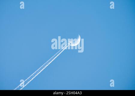 Un avion s'élève haut dans le ciel bleu, laissant des traînées blanches alors qu'il semble traverser la lune, créant une scène aérienne céleste époustouflante. Banque D'Images