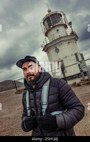 L'homme se tient près d'un vieux phare par une journée nuageuse tout en portant une veste et une casquette, mettant en valeur le paysage côtier accidenté Banque D'Images