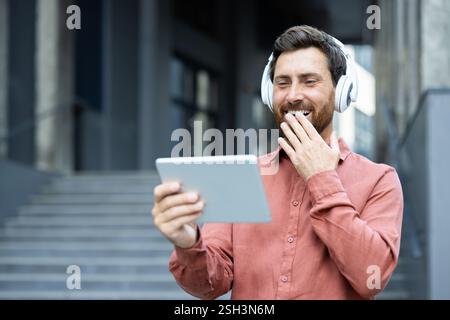 Un homme portant des écouteurs et tenant une tablette regarde un flux vidéo en ligne drôle et divertissant tout en marchant à l'extérieur d'un immeuble de bureaux. Banque D'Images