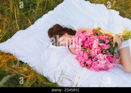Jeune femme dans une robe blanche couchée sur une couverture dans un pré ensoleillé avec un grand bouquet de pivoines roses vibrantes, profitant d'un après-midi d'été paisible. Banque D'Images