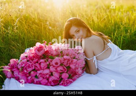 Jeune femme avec un grand bouquet de pivoines roses et dans une robe blanche se trouve sur une couverture sur un arbre dans les rayons du soleil chaud. Idéal pour la publicité, soci Banque D'Images