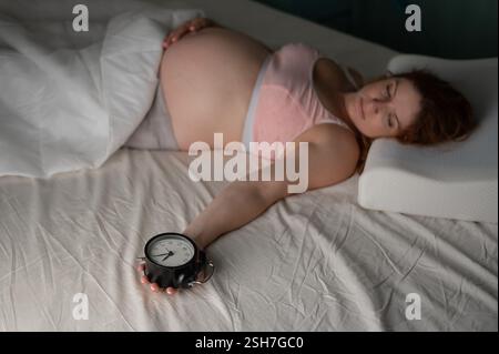 Une femme enceinte se couche dans son lit et tient un réveil. Banque D'Images