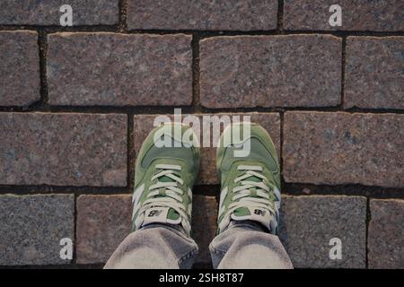 Novembre 2024, Finlande, Helsinki. Vue de dessus, pieds dans les baskets New Balance vertes sur la surface de la rue en briques Banque D'Images