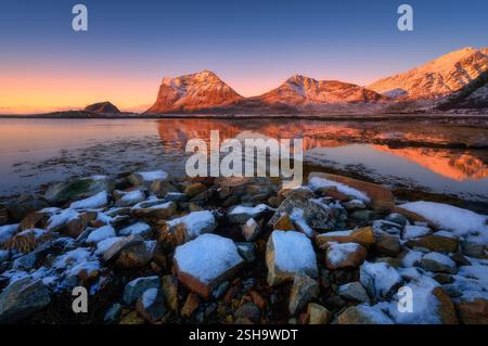 Montagne enneigée, eau calme avec des pierres et ciel coloré au coucher du soleil Banque D'Images
