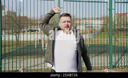 Homme athlétique exécutant des routines d'étirement dynamiques près de la clôture du parc urbain, échauffant les muscles des jambes et des bras avant de courir l'entraînement par jour ensoleillé Banque D'Images