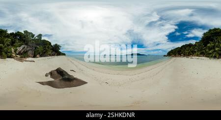 Une vaste étendue de sable blanc fusionne avec la mer tranquille, flanquée d'arbres tropicaux et de formations rocheuses lisses sous un ciel rempli de nuages. La Digue, Seychelles. Vue à 360 degrés. Banque D'Images