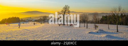 Au coucher du soleil à Prichovice, en Tchéquie, les gens apprécient le paysage hivernal couvert de neige. Les couleurs vibrantes du ciel créent un contraste saisissant avec le terrain blanc et les collines lointaines. Banque D'Images