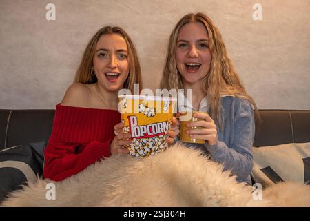 Deux amies joyeuses profitant d'une soirée cinéma sur un canapé confortable, partageant pop-corn et boissons Banque D'Images