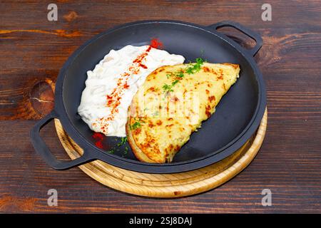 Une omelette dorée est parfaitement cuite et présentée sur une poêle noire, accompagnée d’une sauce riche et crémeuse garnie de persil et d’épices, sertie Banque D'Images