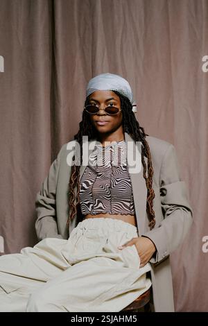 Portrait d'une femme afro-américaine portant une tenue élégante avec un haut à motifs, une veste grise et un pantalon blanc, assise sur une chaise tout en regardant avec confiance la caméra Banque D'Images