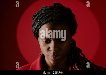 Femme afro-américaine contemplant profondément avec des tresses sur un fond rouge vif. Son regard descendant et son expression paisible véhiculent introspection et sérénité Banque D'Images