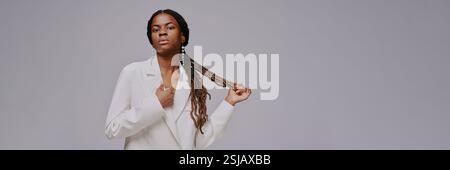 Portrait de femme élégante avec de longues tresses et veste blanche décontractée tenant ses cheveux. L'arrière-plan est minimal et gris, attirant l'attention sur sa position serrée et la texture de ses tresses Banque D'Images