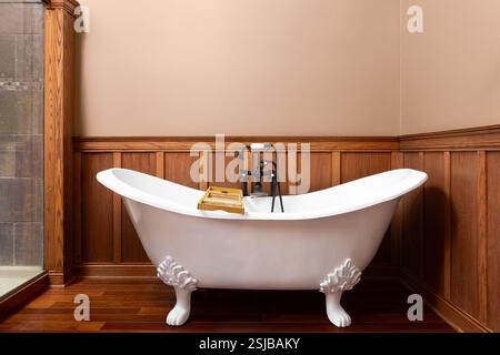 La salle de bains est dotée d'une baignoire sur pieds à griffes dotée de murs lambrissés et d'un parquet en bois franc, de murs peints en brun et d'une douche carrelée en pierre. Banque D'Images