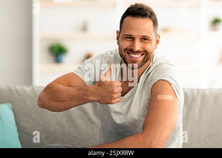 Un gars heureux approuvant les vaccinations, montrant du plâtre médical sur l'épaule après l'injection et en faisant des gestes de pouce vers le haut, espace libre Banque D'Images