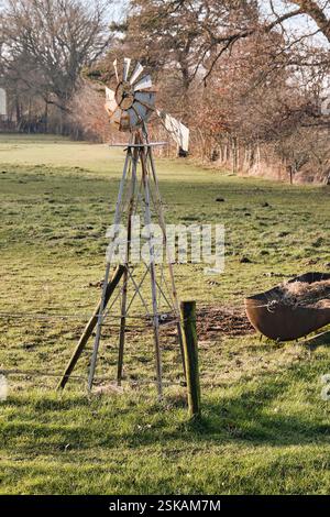 Un moulin à vent altéré se dresse dans un pâturage herbeux, entouré d'arbres et d'une clôture rustique, capturant le charme de la campagne en plein jour. Banque D'Images