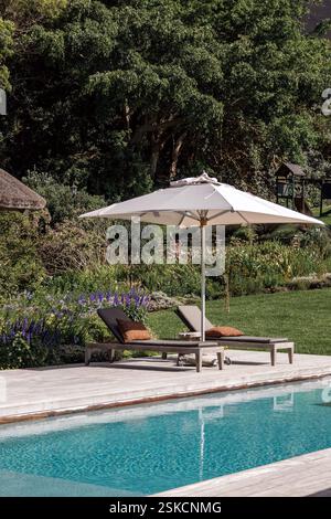 un parasol blanc jette une ombre sur deux chaises longues rembourrées près de la piscine. En arrière-plan, un jardin luxuriant et verdoyant, avec diverses plantes sp Banque D'Images