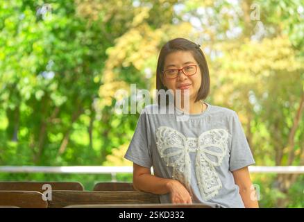 Portrait de femme asiatique d'âge moyen dans l'humeur positive avec le fond d'arbres flous, la lumière naturelle du soleil, femme asiatique en bonne santé portant des lunettes, s. Banque D'Images