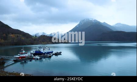 Un petit quai flottant sur un côté de la photo, dans un fjord norvégien calme, plusieurs bateaux sont amarrés. Au centre, des montagnes enneigées. Banque D'Images