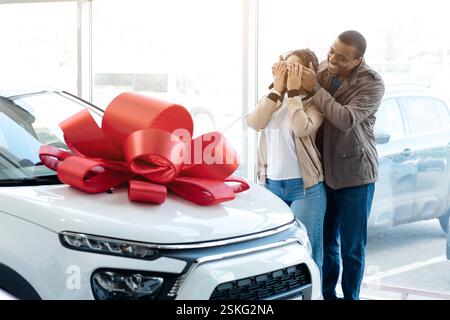 Propre voiture. Mari amrericain africain couvrant les yeux de sa femme montrant sa nouvelle auto dans le magasin de la concession. Espace vide Banque D'Images