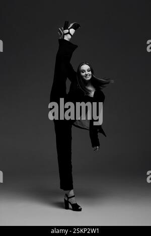 Danseuse féminine flexible dans une tenue noire élégante exécutant une extension de jambe haute dans un studio. Parfait pour la danse, le fitness ou les concepts artistiques. Banque D'Images