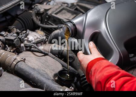 Gros plan de la main d'un mécanicien versant de l'huile moteur fraîche d'un grand récipient en plastique dans le bouchon de remplissage d'huile d'un moteur de voiture. Le travailleur, habillé Banque D'Images