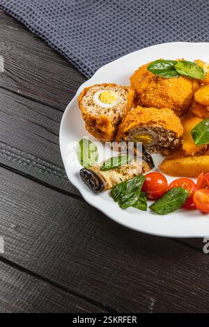 Une assiette présente des roulés de poulet doré farcis d'oeufs, accompagnés de pommes de terre rôties croustillantes, de tomates cerises et de feuilles de basilic frais, sertis Banque D'Images