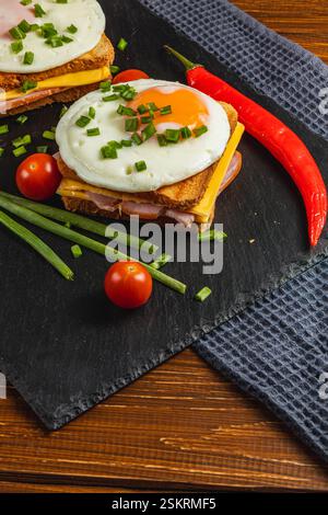 Un sandwich copieux pour le petit-déjeuner comprend des couches de fromage, de jambon et de pain grillé croustillant surmonté d'œufs frits, de tomates cerises et d'herbes fraîches, le tout sur un plan de d. Banque D'Images