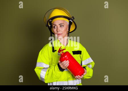 Pompier femme mature dans un uniforme de protection lumineux tenant un extincteur rouge, placé sur un fond kaki symbolisant le sauvetage Banque D'Images