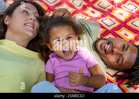 Heureuse famille mixte allongée sur une couverture de pique-nique colorée riant ensemble à l'extérieur en été Banque D'Images