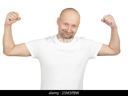 Un homme chauve caucasien portant un t-shirt blanc fléchit ses bras dans une pose confiante. Il a un léger sourire sur son visage, exprimant positivité et str Banque D'Images