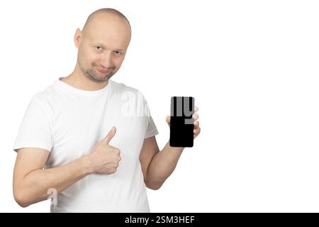 Un homme caucasien chauve avec une barbe courte tient un smartphone dans une main et montre un pouce vers le haut avec l'autre. Banque D'Images