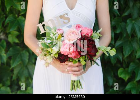 Une mariée tient un bouquet vibrant avec des roses roses et des pivoines, avec un signe Mme, debout au milieu d'un feuillage vert luxuriant. Banque D'Images