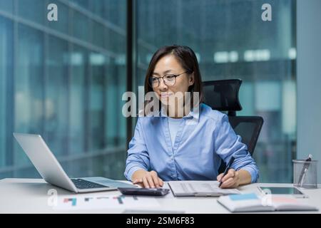 Sérieuse femme asiatique focalisée faisant de la paperasse à l'intérieur du bureau. Femme d'affaires remplissant des formulaires fiscaux et des rapports financiers. Femme réfléchie utilisant un ordinateur portable au travail sur le lieu de travail. Banque D'Images