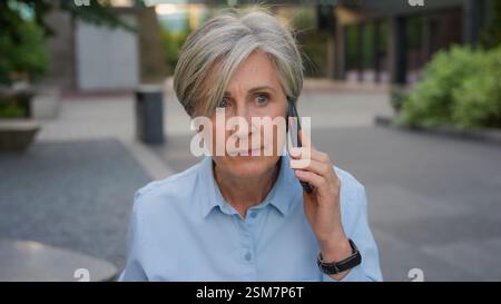 En colère folle femme d'affaires senior caucasienne vieille femme stressée femme argumente parler téléphone portable mécontent dame d'affaires mature sur la rue de la ville parler Banque D'Images