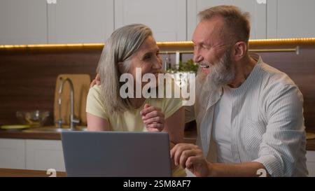 Heureux 60s Caucasien femme mariée femme et homme mari parlant discuter des réseaux sociaux riant shopping en ligne avec ordinateur portable dans la cuisine Internet Banque D'Images