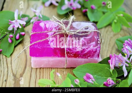 Deux barres de savon rose maison, nouées avec de la ficelle, des brindilles avec des feuilles et des fleurs roses de chèvrefeuille sur un fond de planches de bois Banque D'Images