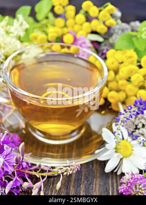 Tisane dans une tasse en verre à partir de fleurs fraîches d'épilobes, tanaisie, camomille, trèfle, millefeuille, la reine-des-Prés, les feuilles de menthe sur un fond en bois Banque D'Images