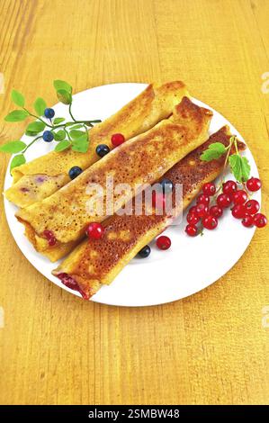 Crêpes avec remplissage de baies sur une assiette en porcelaine blanche, deux pinceaux de groseille rouge et brindille de myrtille avec des feuilles vertes sur un fond de bois Banque D'Images