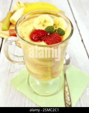 Lait de dessert avec du lait caillé, banane, fraises et rhubarbe, cuillère sur une serviette en papier vert sur le fond des planches en bois clair Banque D'Images