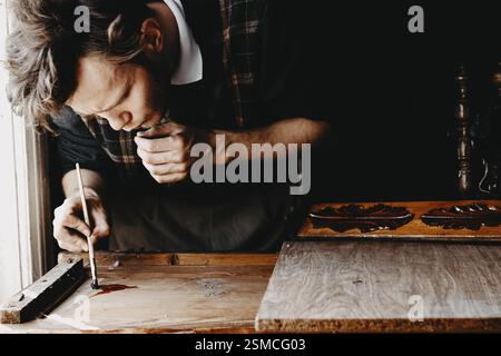 Un homme concentré travaillant avec un pinceau sur du bois près d'une fenêtre Banque D'Images